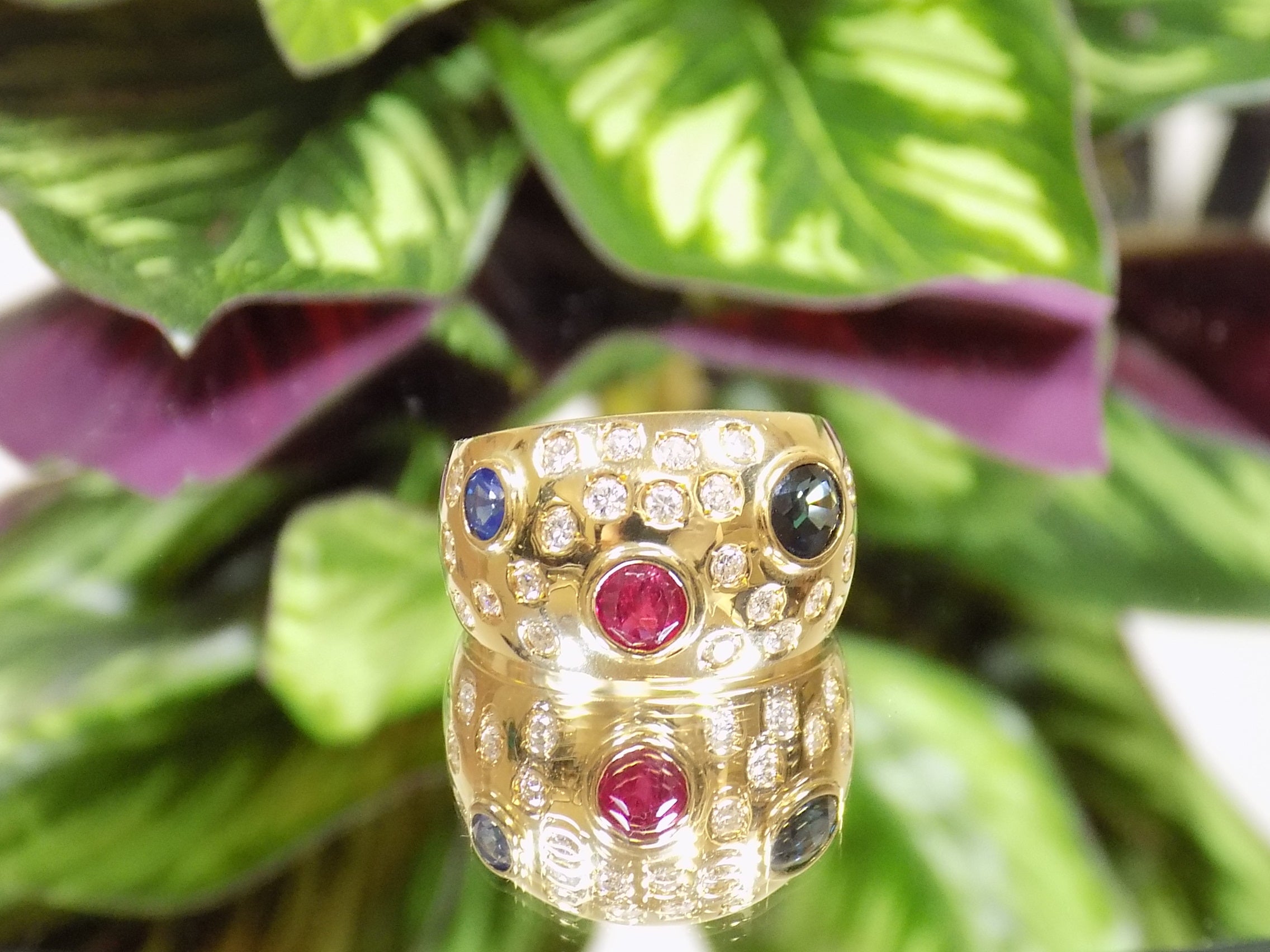 This is a women's estate ring with 2.50ct total weight ruby, sapphire, and diamonds set in 18K yellow gold on a mirrored surface with a green leafy background.