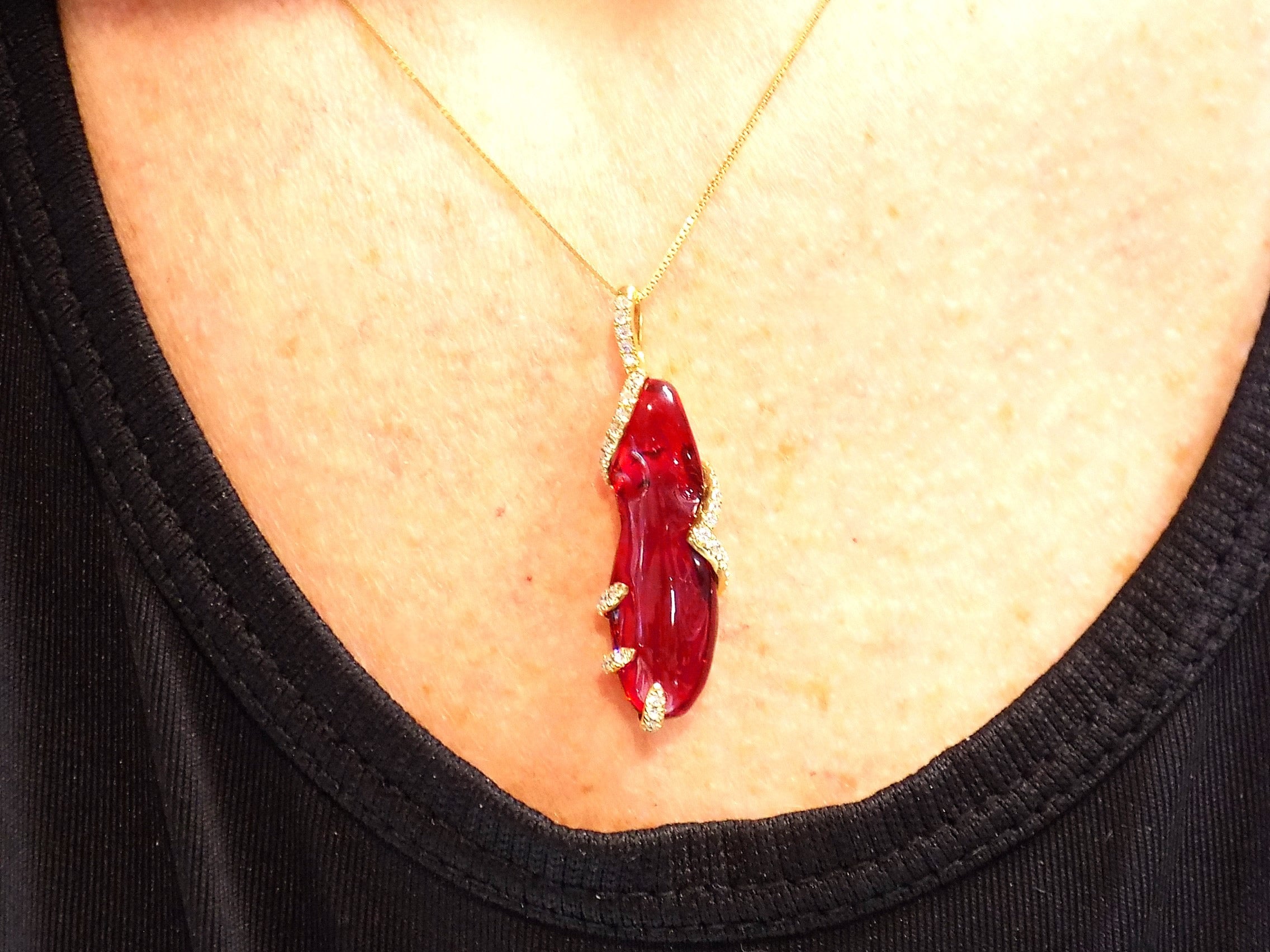 This is a women's custom made necklace with a 6.63ct red fire opal uncut but polished in a custom built 14K yellow gold mounting with .25ct round diamonds making it a 6.88ct total weight on a women's neck wearing a black shirt.