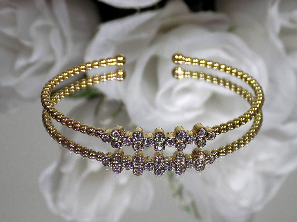 This is a .50ct natural round brilliant diamond flex bangle in 10K yellow gold on a mirrored surface with a white floral background.