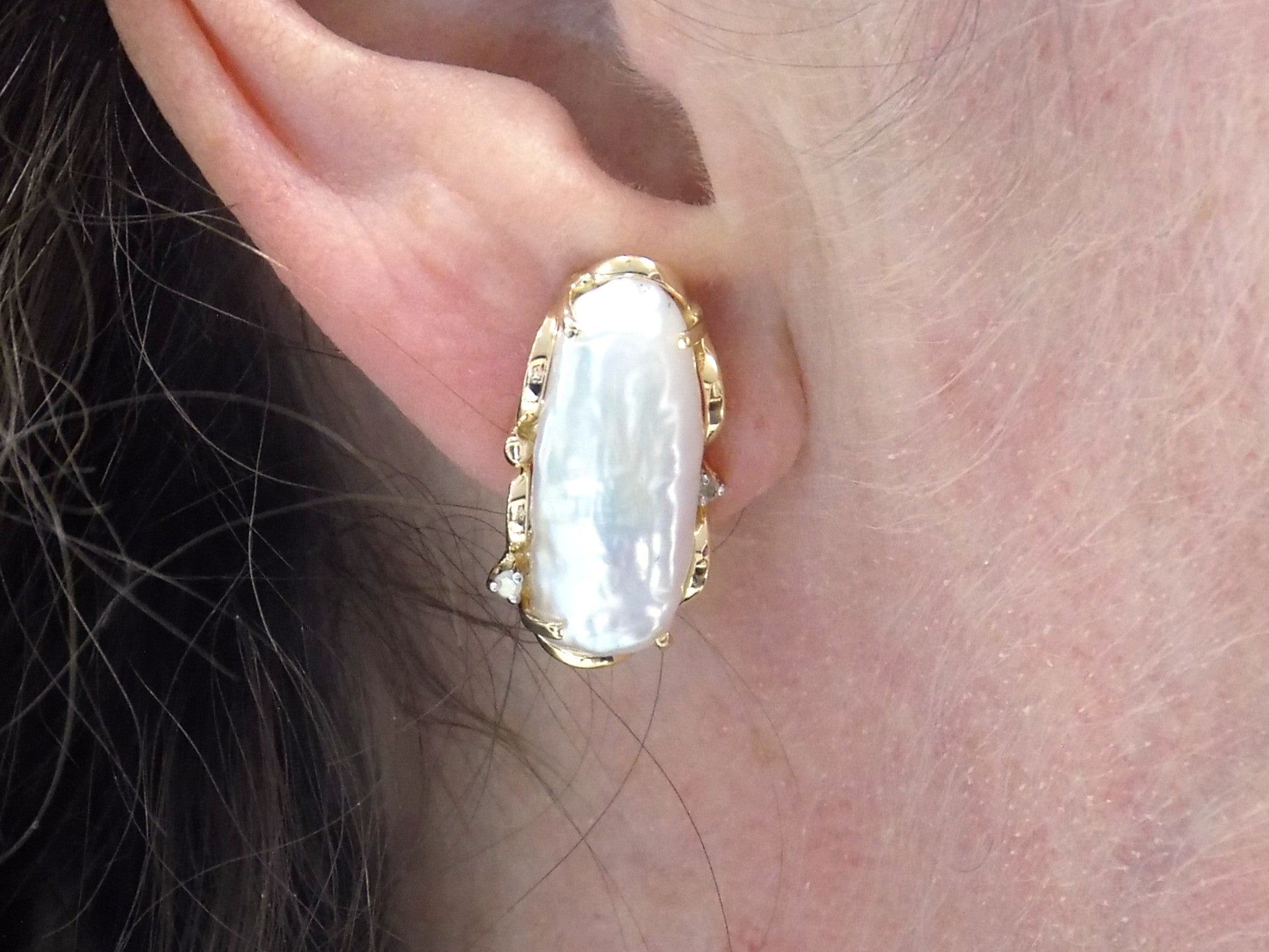These are genuine large elongated freshwater pearl earrings with omega backs in yellow gold displayed in a woman's ear.