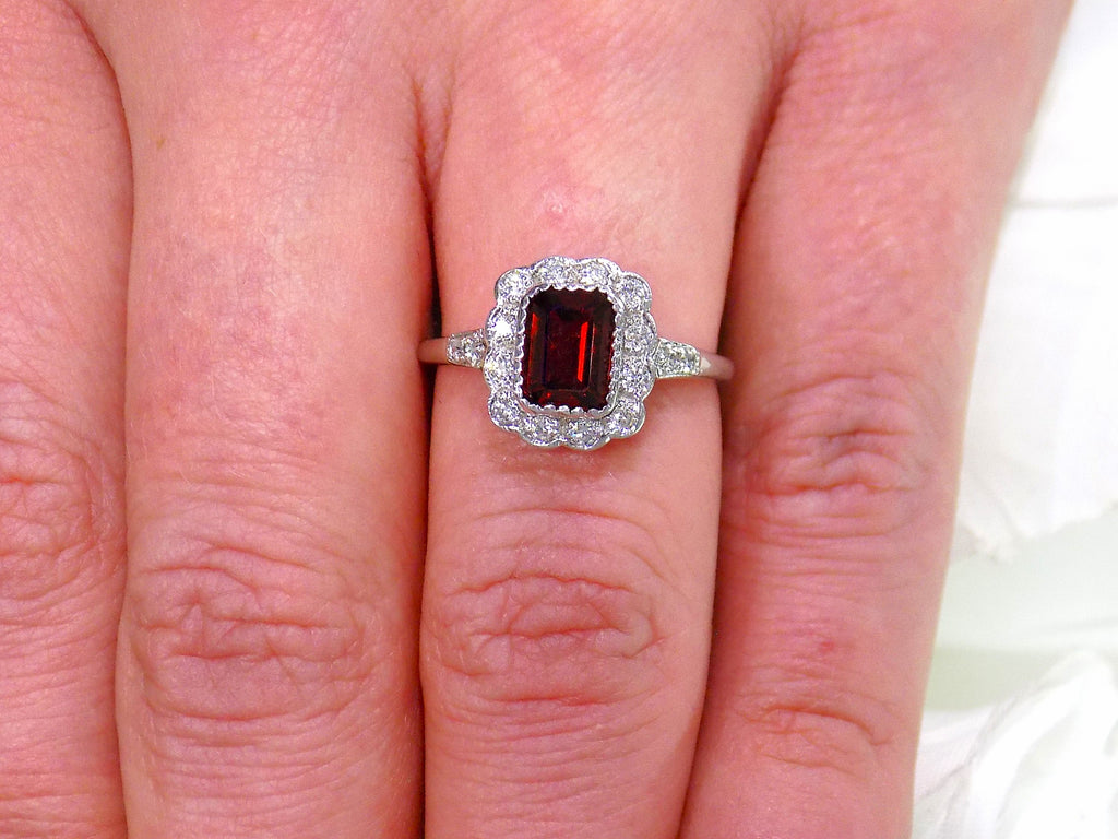 This is an emerald cut genuine garnet ring surrounded by natural diamonds in white gold and fully restored to like new shown on a ladies' finger.