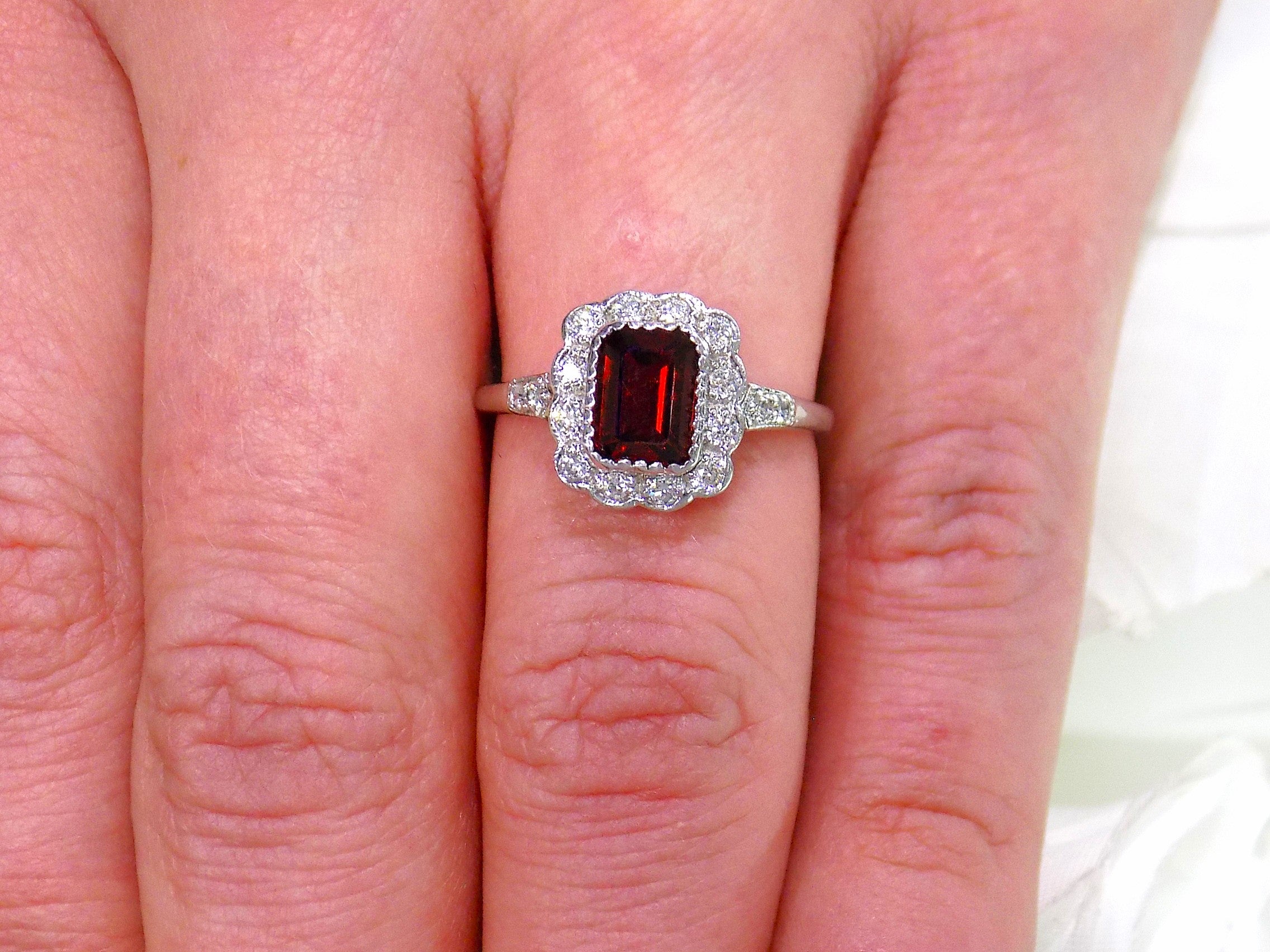 This is an emerald cut genuine garnet ring surrounded by natural diamonds in white gold and fully restored to like new shown on a ladies' finger.