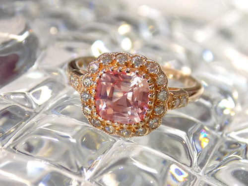 This is a women's ring with 1.94ct natural cushion cut morganite in the center surrounded by .30ct in natural round diamond accents in 14K rose gold sitting on a textured glass surface.