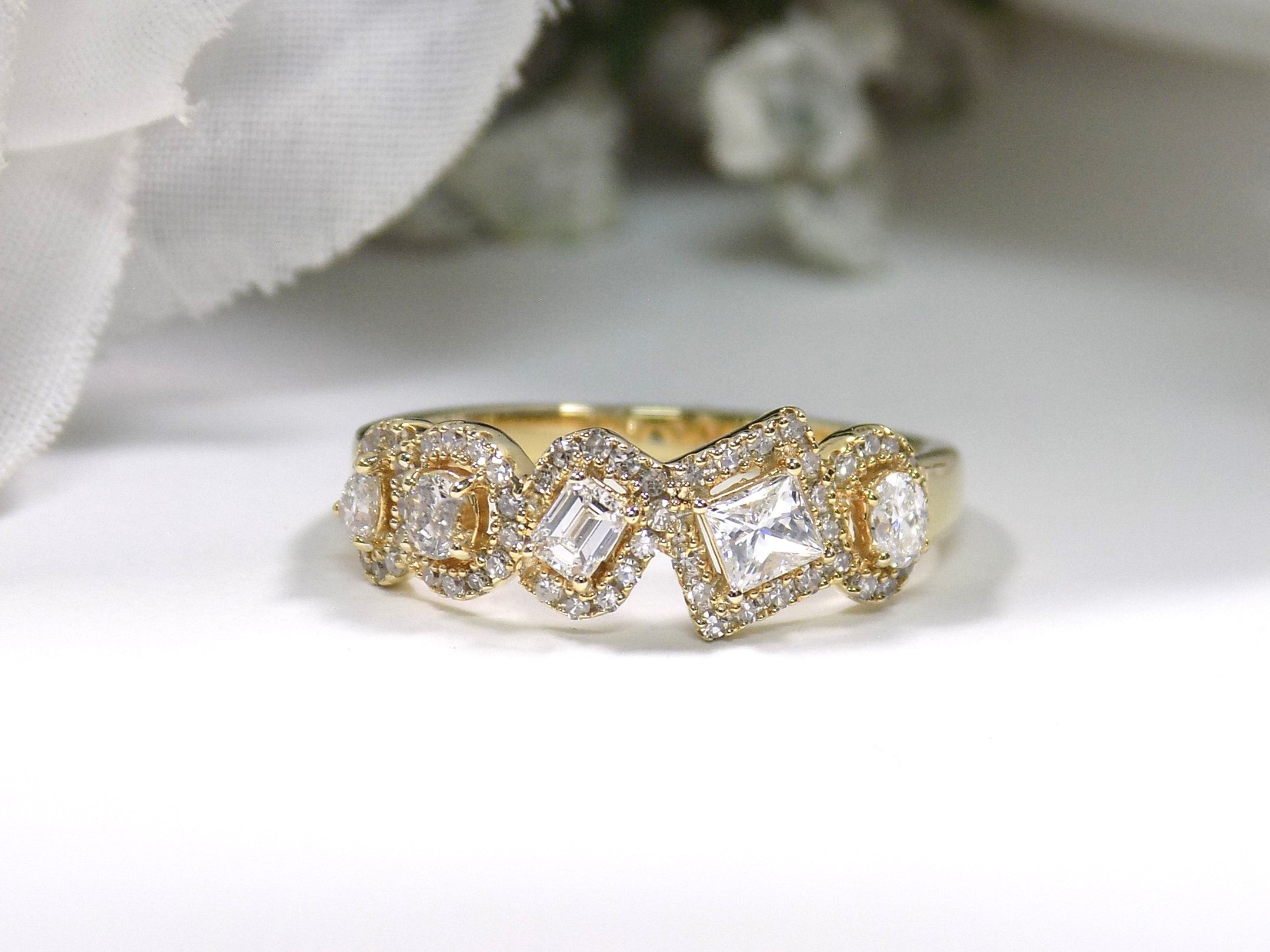 This is a multi shape natural diamond ring including a pea shape, round brilliant, emerald cut, princess cut and oval shape all surrounded by round diamond accents in 10K yellow gold on a white background.