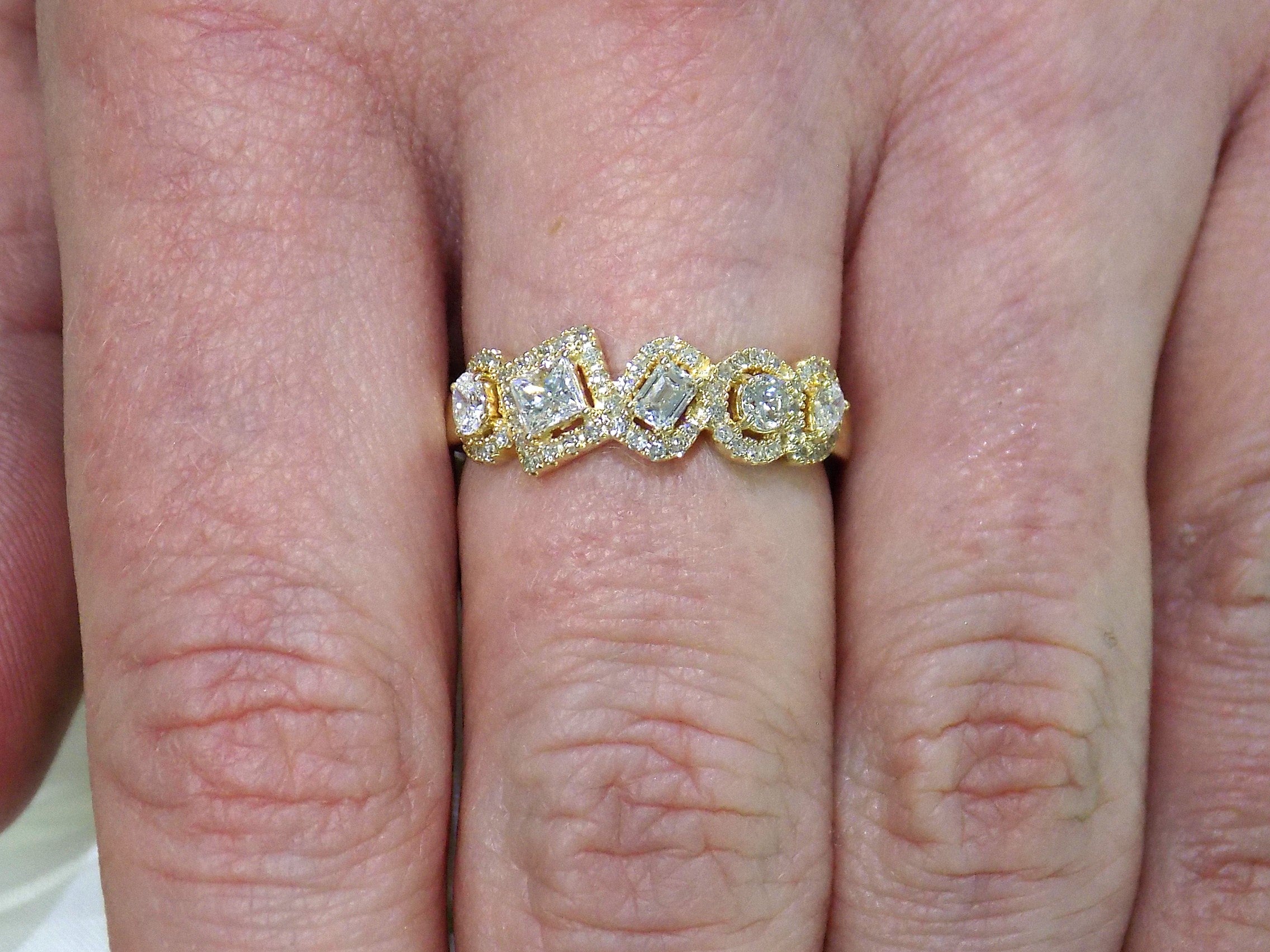 This is a multi shape natural diamond ring including a pea shape, round brilliant, emerald cut, princess cut and oval shape all surrounded by round diamond accents in 10K yellow gold on a ladies finger.