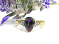 This is a women's ring with a 2.00ct pear shape mystic fire topaz center in green and purple with .12ct natural accent diamonds set in 14K yellow gold on a white surface with a purple and green floral background.