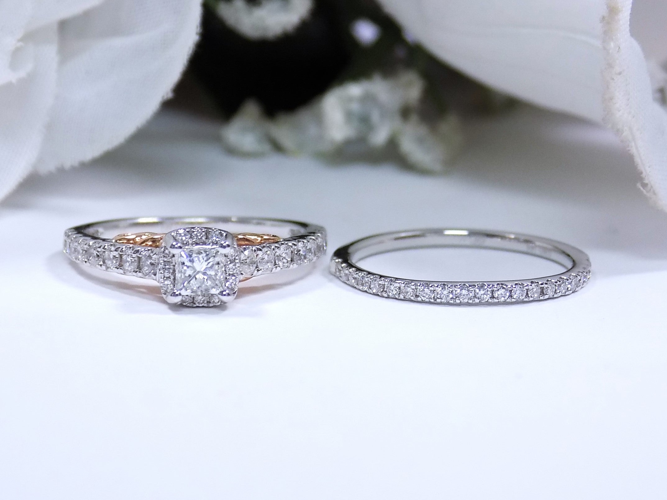 This is a halo style women's 2-piece wedding set including the engagement ring and matching band laying side by side with a princess cut center weighing .88ct total in white gold with rose accents shown with a white background.