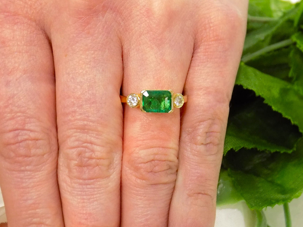 This is a woman's natural emerald cut emerald split bezel set sideways with a sing bezel set round diamond on either side in yellow gold shown on a woman's finger.