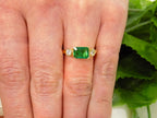 This is a woman's natural emerald cut emerald split bezel set sideways with a sing bezel set round diamond on either side in yellow gold shown on a woman's finger.