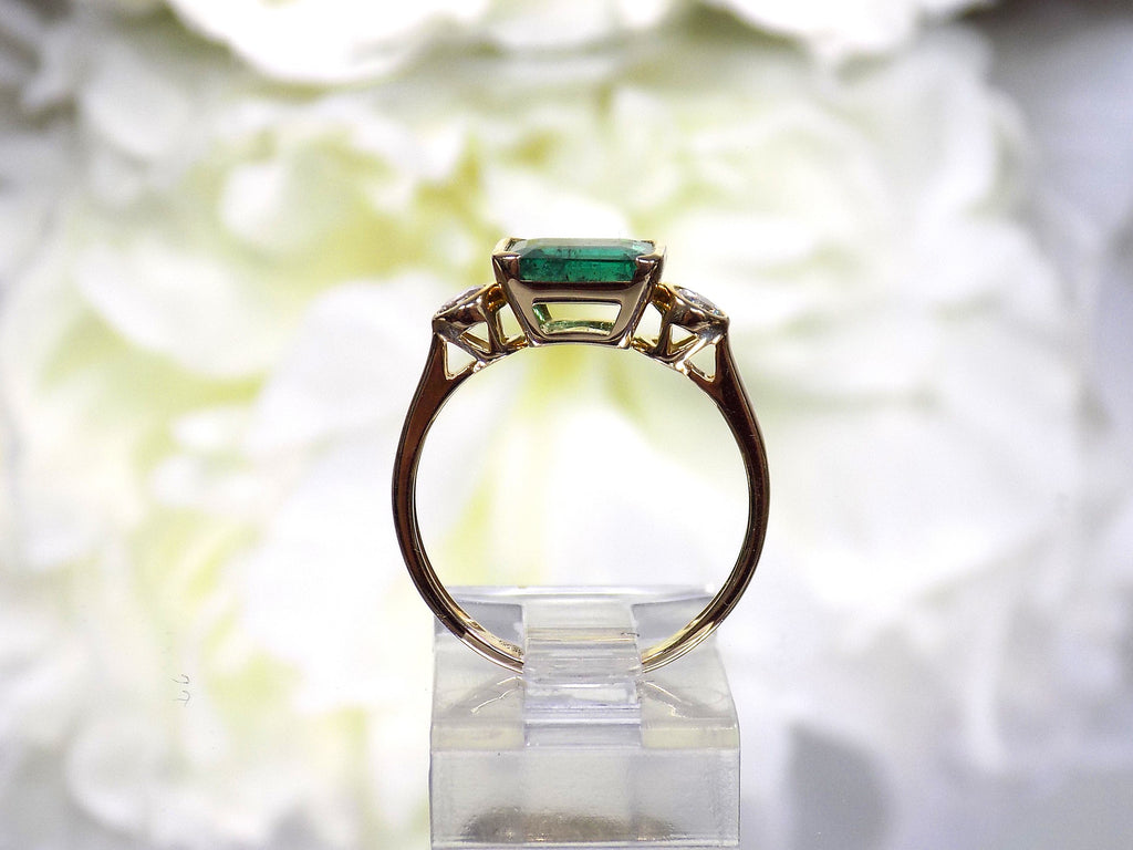 This is a side view of woman's natural emerald cut emerald split bezel set sideways with a sing bezel set round diamond on either side in yellow gold shown with a white flowery background.