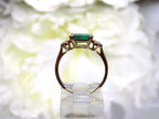 This is a side view of woman's natural emerald cut emerald split bezel set sideways with a sing bezel set round diamond on either side in yellow gold shown with a white flowery background.