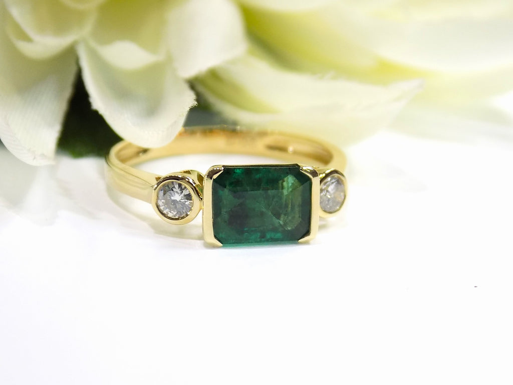 This is a woman's natural emerald cut emerald split bezel set sideways with a sing bezel set round diamond on either side in yellow gold shown with a white flowery background.