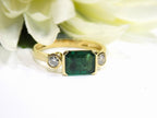 This is a woman's natural emerald cut emerald split bezel set sideways with a sing bezel set round diamond on either side in yellow gold shown with a white flowery background.