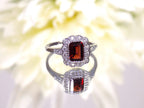 This is a women's with a natural 1.00ct bezel set emerald cut garnet surrounded by a single row of .28ct total weight natural diamond accents set in 10K white gold on a mirrored surface with a floral background.
