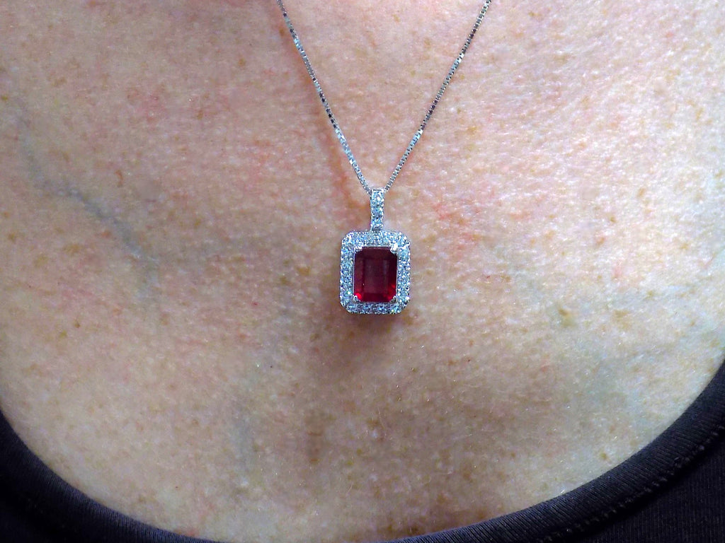 This is a women's pendant 3.65ct natural enhanced emerald cut ruby center surrounded by 1.56ct total weight in natural round diamond accents in 14K white gold on a women's neck with a black shirt.
