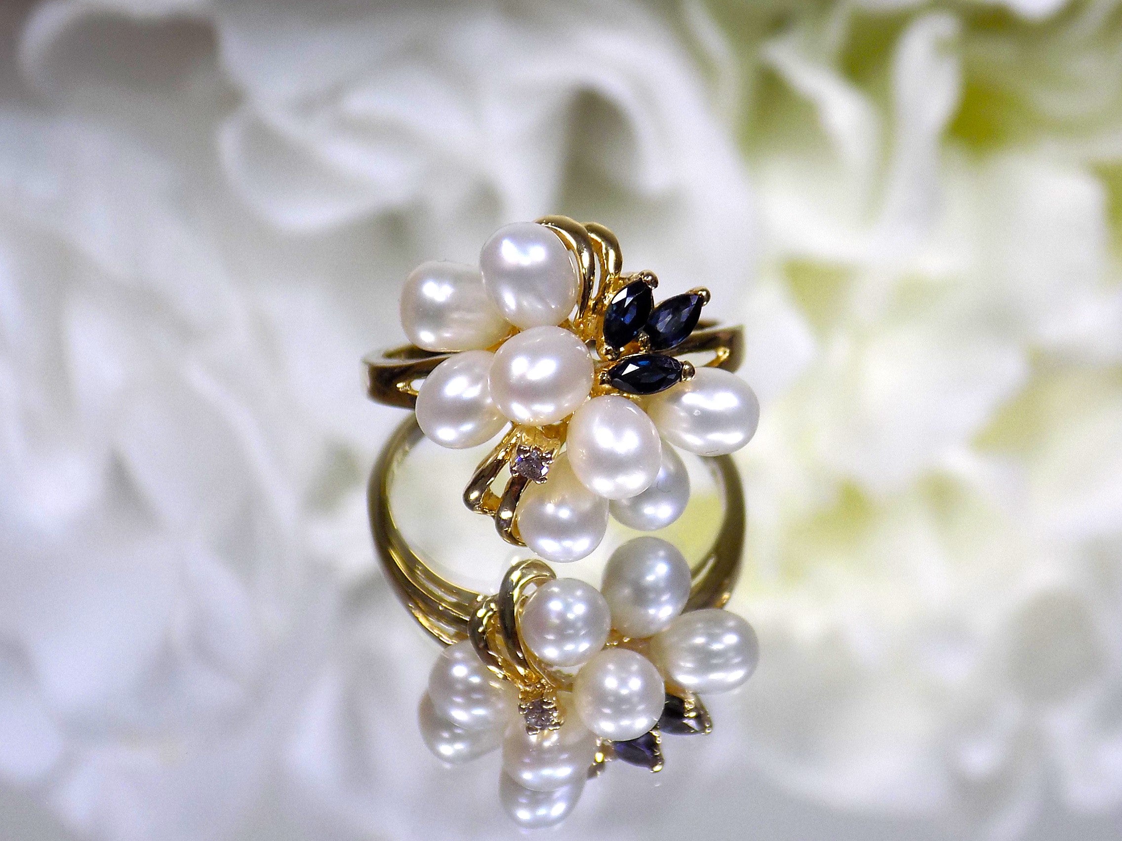 This is a freshwater pearl, blue sapphire, and diamond ring in yellow gold from our estate collection with a mirrored flower background.