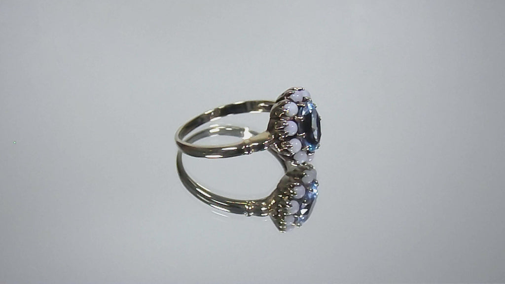 This a video of a ring with an oval blue topaz in the center and surrounded by natural opals in yellow gold from our estate collection turning slowly on mirrored background.