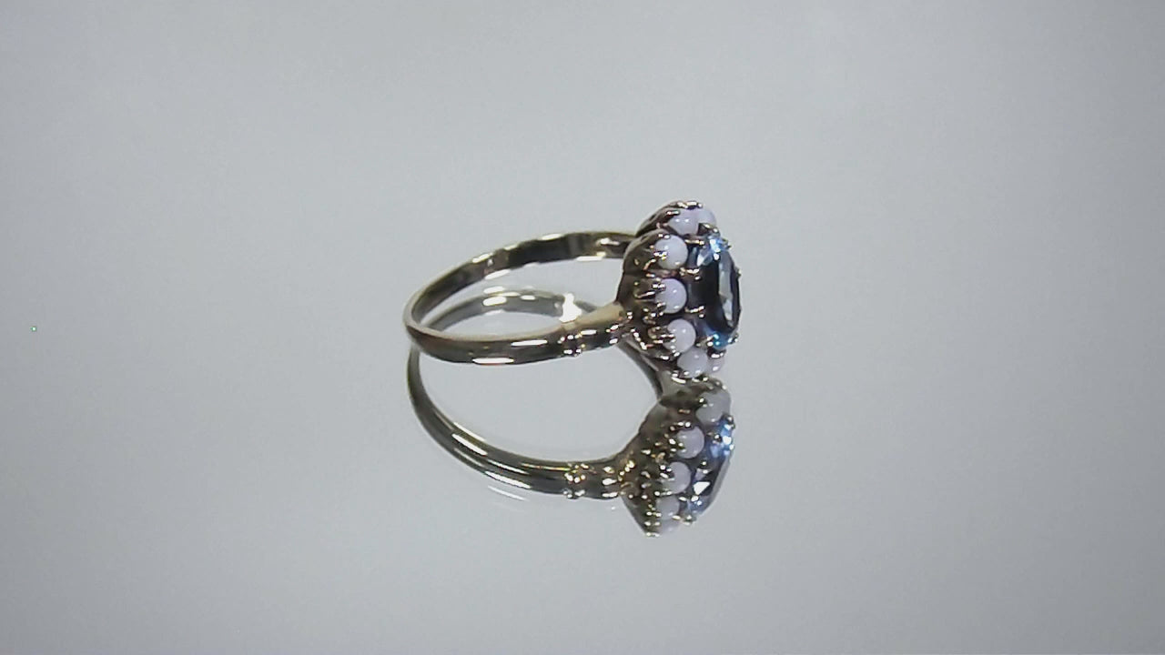 This a video of a ring with an oval blue topaz in the center and surrounded by natural opals in yellow gold from our estate collection turning slowly on mirrored background.