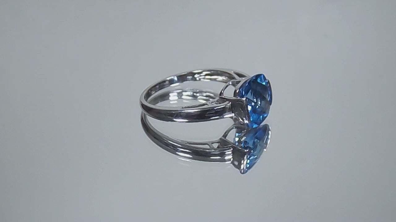 This is a tension set or channel set 4.00ct natural cushion cut blue topaz solitaire ring in 14K white gold on a mirrored surface rotating in a video.