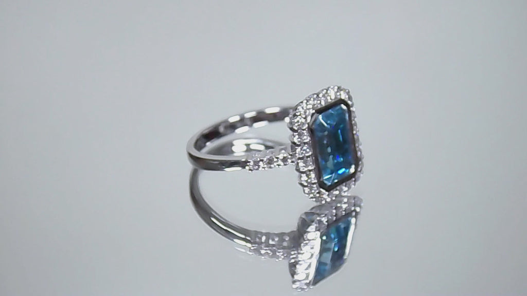 This is a women's ring has a 3.35ct natural emerald cut blue zircon in the center with .49ct natural round diamond accent's on a mirrored background rotating in a video.
