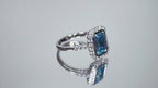 This is a women's ring has a 3.35ct natural emerald cut blue zircon in the center with .49ct natural round diamond accent's on a mirrored background rotating in a video.