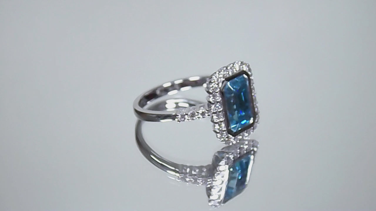 This is a women's ring has a 3.35ct natural emerald cut blue zircon in the center with .49ct natural round diamond accent's on a mirrored background rotating in a video.