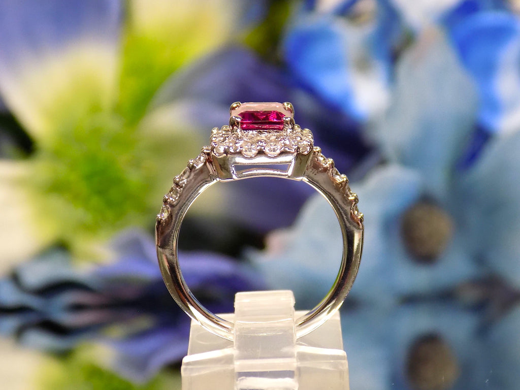 This is a women's ring with a 1.47ct natural emerald cut pink tourmaline surrounded by .67ct natural round diamonds set in 14K white gold on a mirrored surface with a blue floral background from a side view.