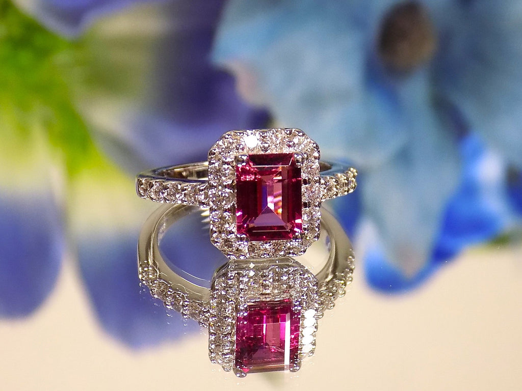 This is a women's ring with a 1.47ct natural emerald cut pink tourmaline surrounded by .67ct natural round diamonds set in 14K white gold on a mirrored surface with a blue floral background.