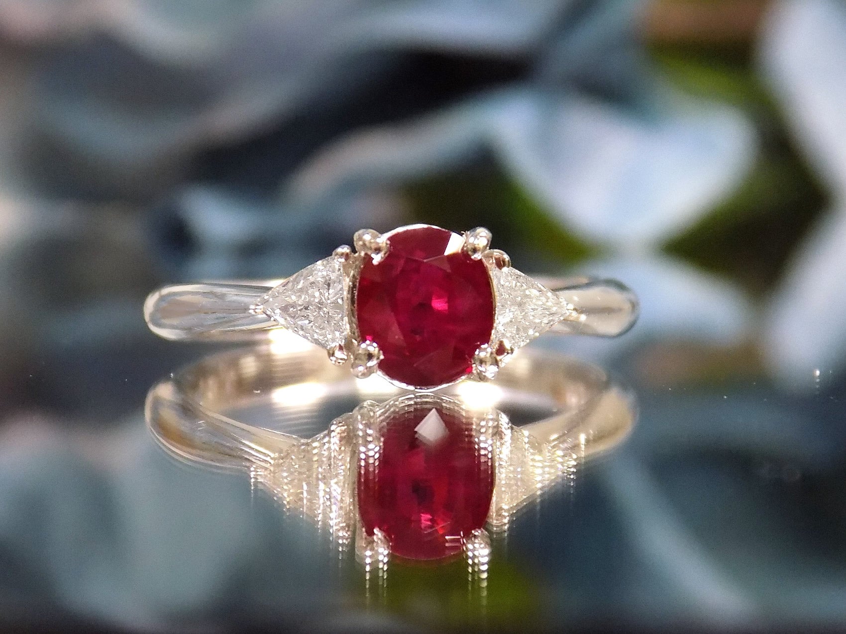 This a 1.51ct total weight natural oval pigeon blood red ruby with a single trillion shaped natural diamond on either side in platinum from our estate collection with a flowery mirrored background. 