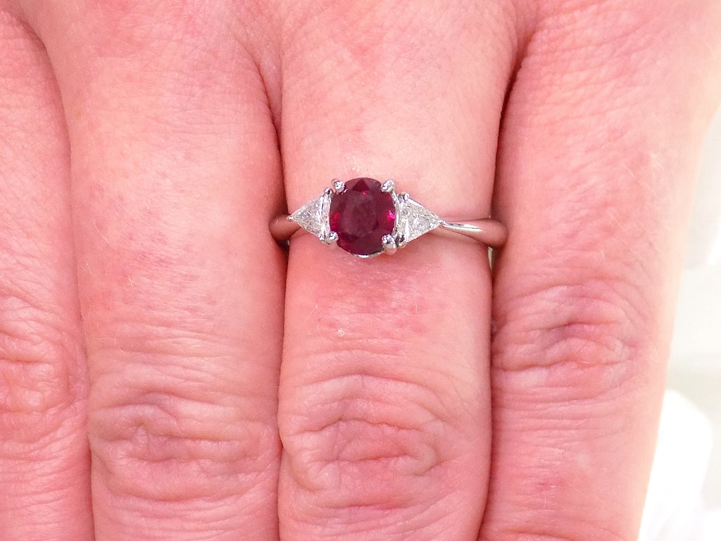 This a 1.51ct total weight natural oval pigeon blood red ruby with a single trillion shaped natural diamond on either side in platinum from our estate collection shown on a woman's finger.