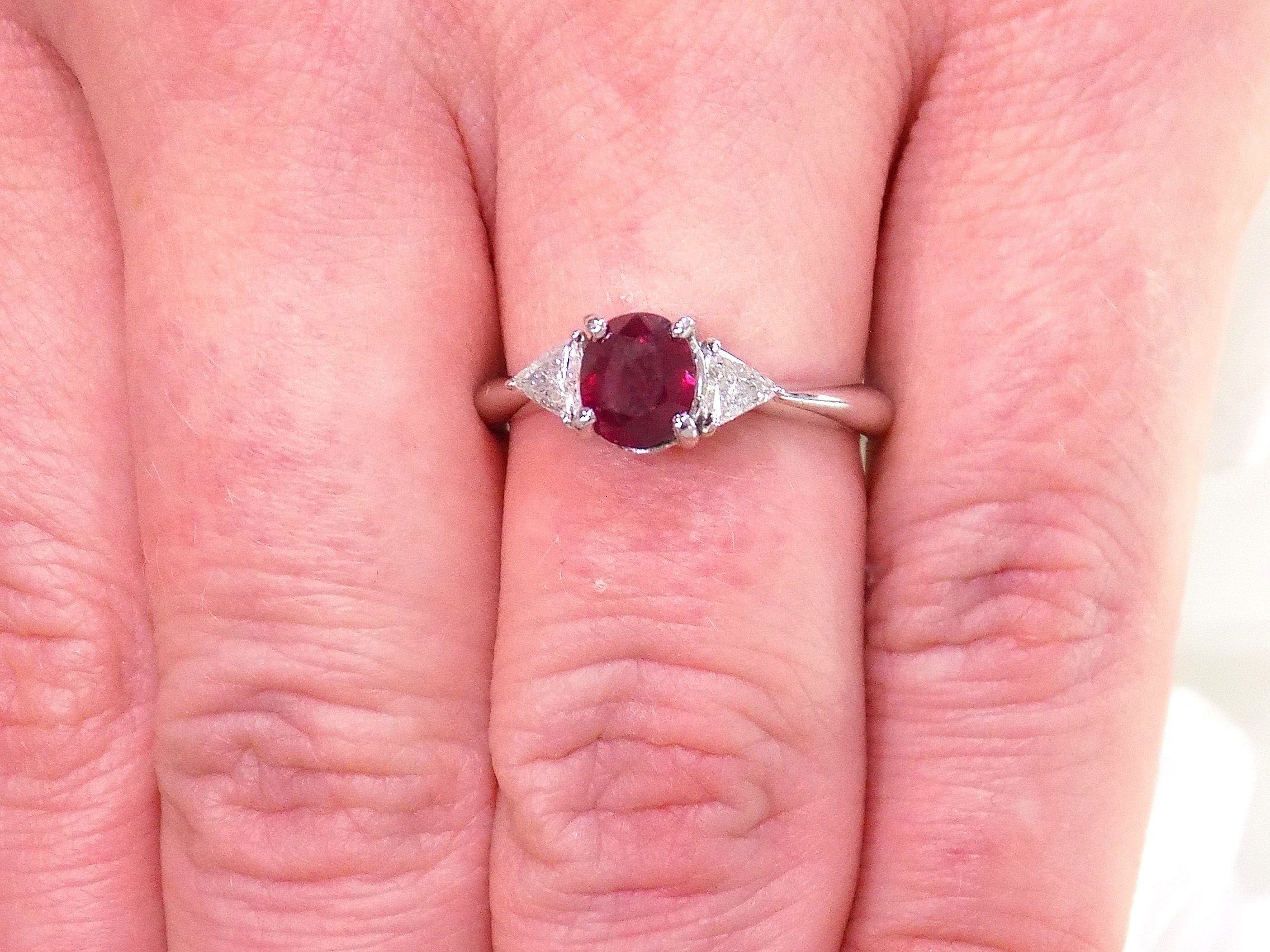 This a 1.51ct total weight natural oval pigeon blood red ruby with a single trillion shaped natural diamond on either side in platinum from our estate collection shown on a woman's finger.