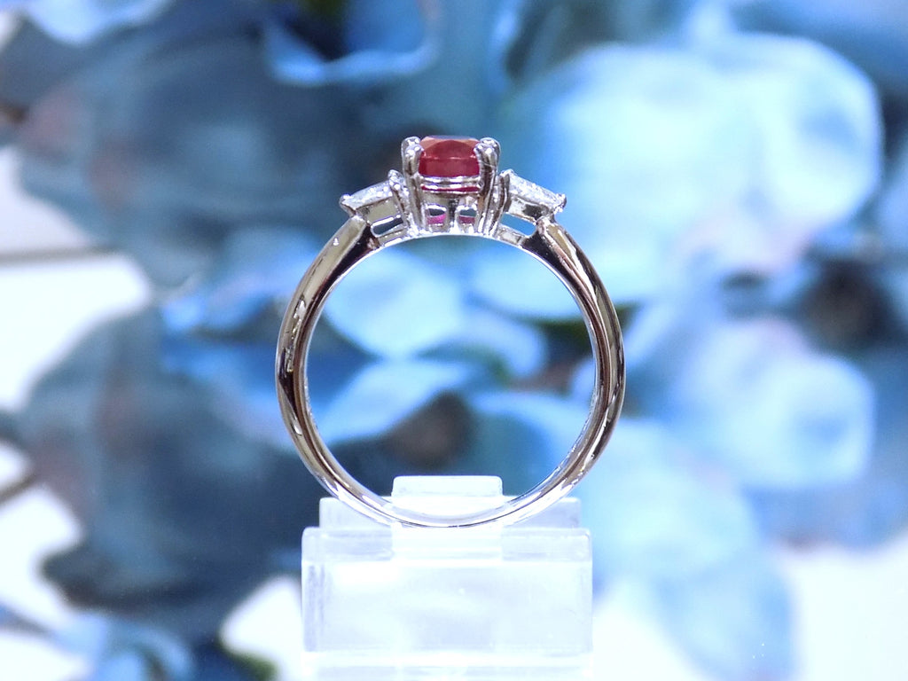 This a side view of a 1.51ct total weight natural oval pigeon blood red ruby with a single trillion shaped natural diamond on either side in platinum from our estate collection with a blue flowery mirrored background.