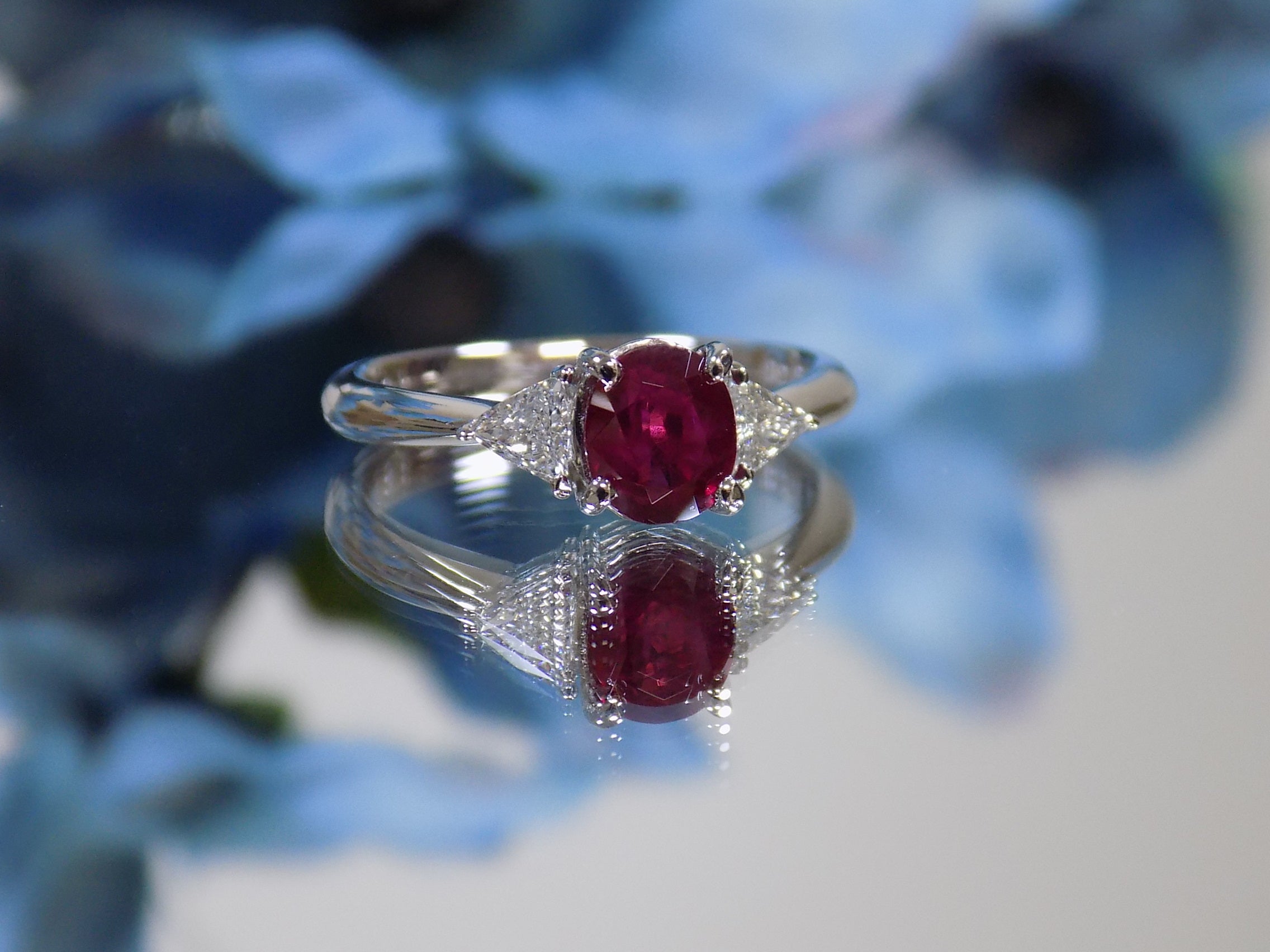 This a 1.51ct total weight natural oval pigeon blood red ruby with a single trillion shaped natural diamond on either side in platinum from our estate collection with a blue flowery mirrored background. 