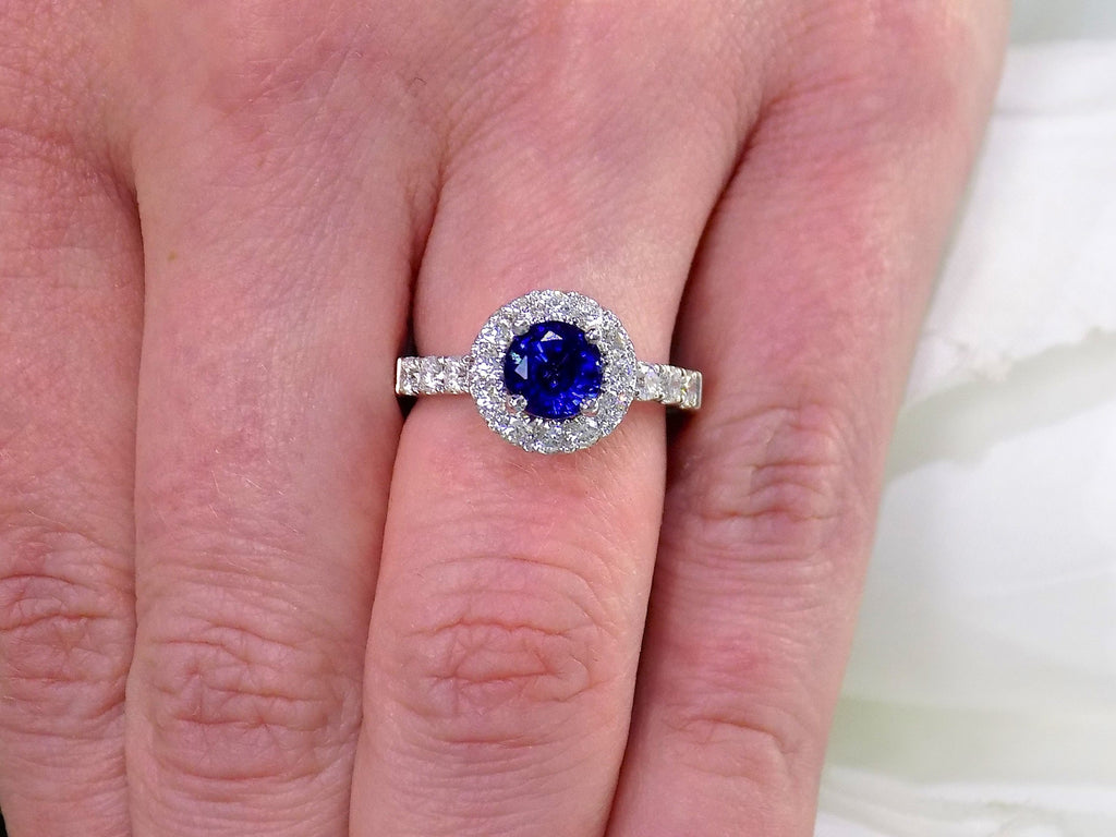 This is a women's halo style 2ct total weight round sapphire and diamond ring in white gold shown on a woman's finger.