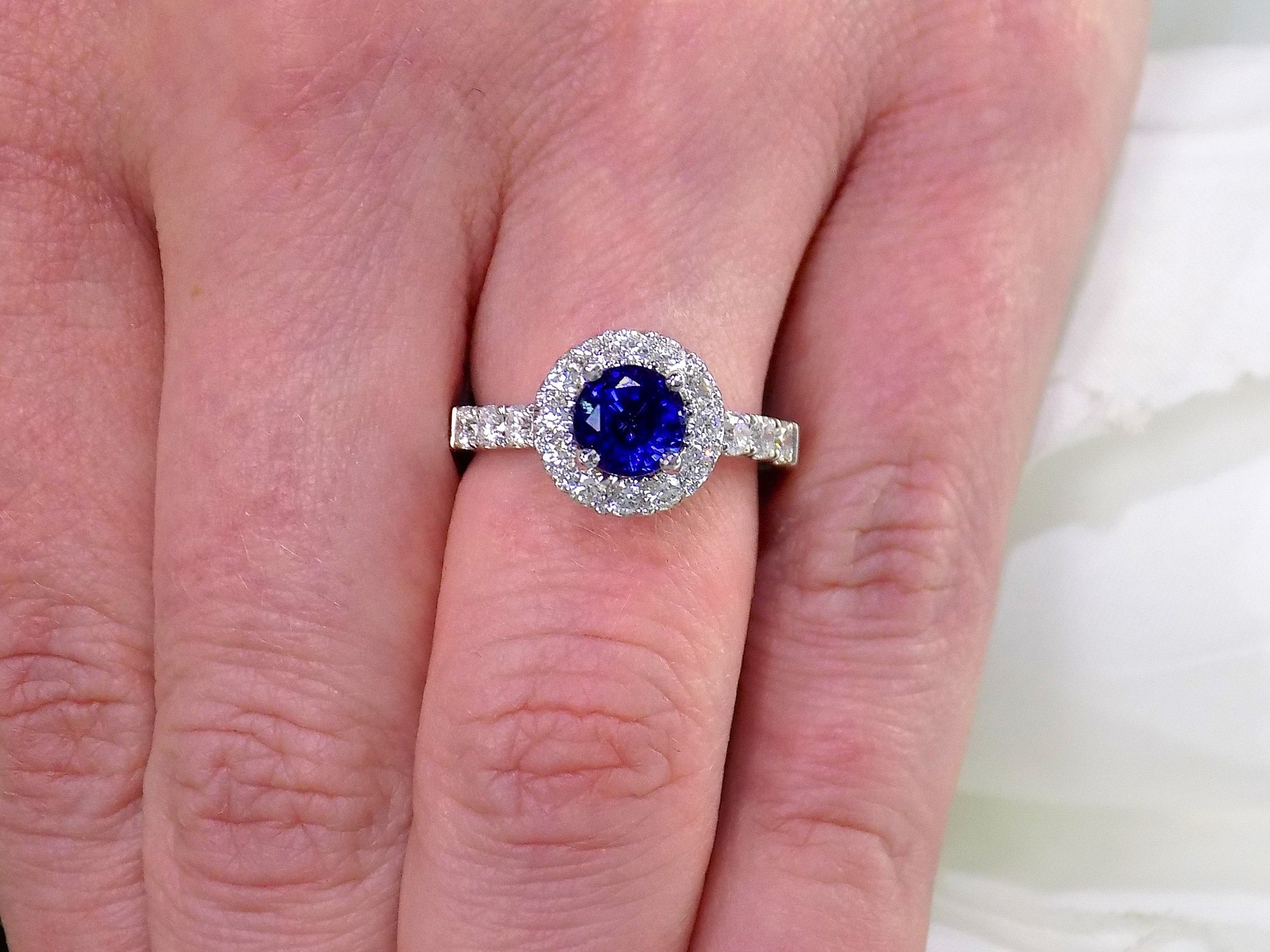 This is a women's halo style 2ct total weight round sapphire and diamond ring in white gold shown on a woman's finger.