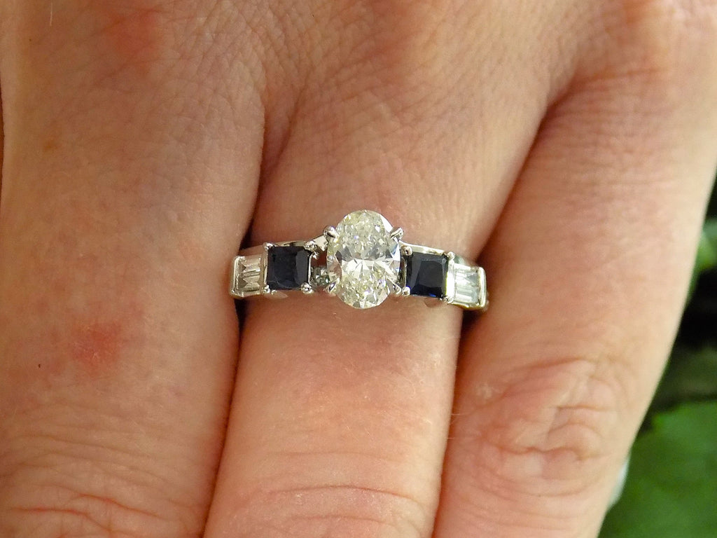 This is a white gold sapphire and diamond ring guard or wrap with an oval solitaire ring in the center shown on a ladies' finger.
