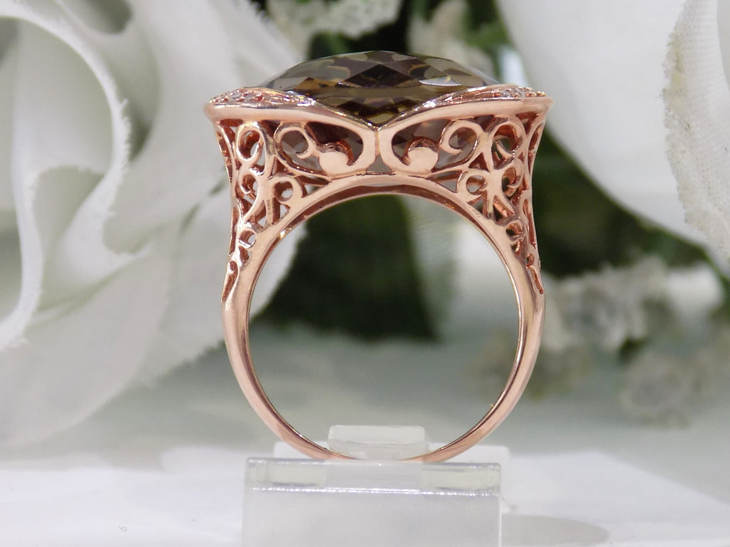 This is a women's ring with a fantasy cut approximate 13ct natural smoky quartz or topaz in the center with .08ct in natural diamonds on both sides set in a filigree style 14K rose gold mounting with a white floral background from a side view of the ring.
