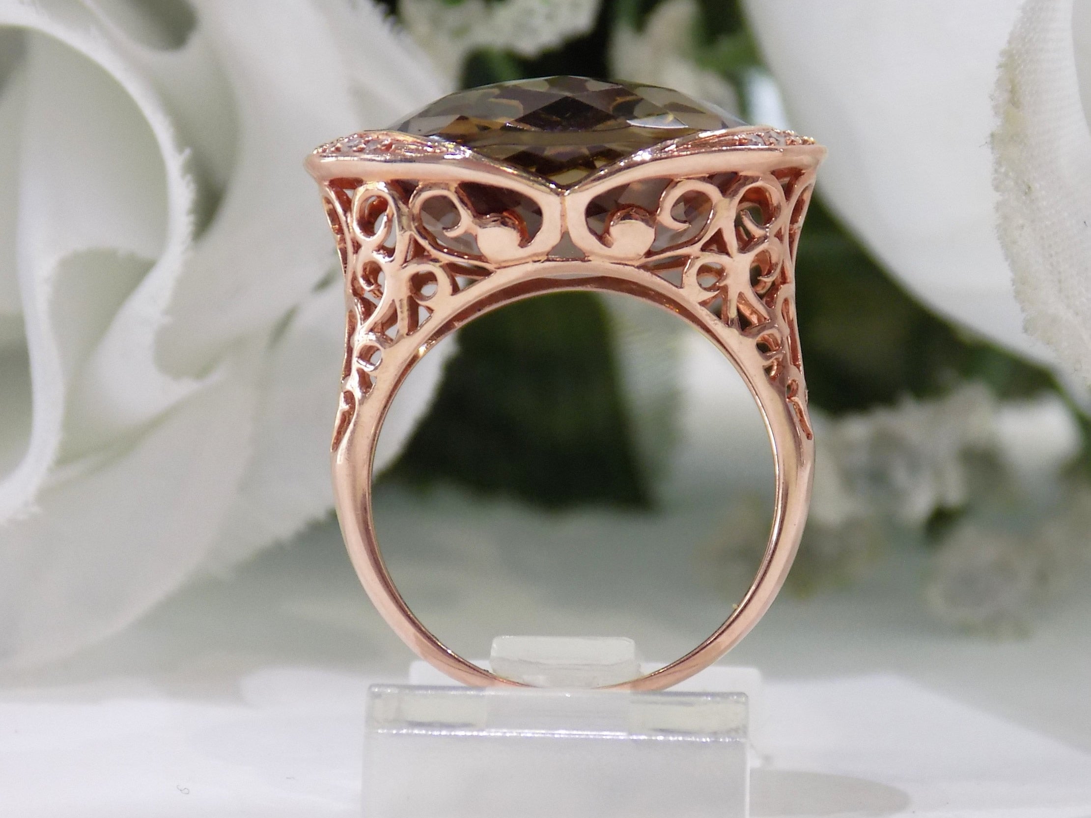 This is a women's ring with a fantasy cut approximate 13ct natural smoky quartz or topaz in the center with .08ct in natural diamonds on both sides set in a filigree style 14K rose gold mounting with a white floral background from a side view of the ring.