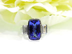 This is a 4.50ct total weight natural bezel set elongated cushion cut tanzanite ring with a single bezel set natural diamond on either side shown yellow flowery background.