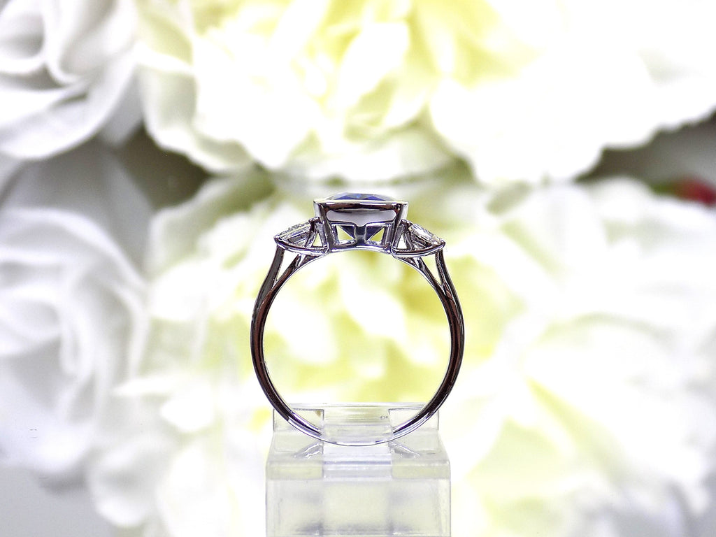 This is a side view of a 4.50ct total weight natural bezel set elongated cushion cut tanzanite ring with a single bezel set natural diamond on either side shown with a white flowery background.
