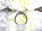 This is a side view of a 4.50ct total weight natural bezel set elongated cushion cut tanzanite ring with a single bezel set natural diamond on either side shown with a white flowery background.