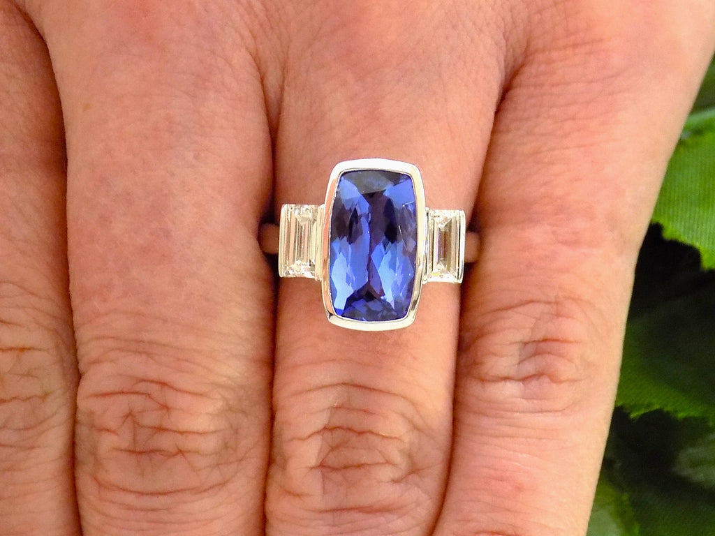 This is a 4.50ct total weight natural bezel set elongated cushion cut tanzanite ring with a single bezel set natural diamond on either side shown on a woman's finger.