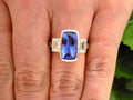 This is a 4.50ct total weight natural bezel set elongated cushion cut tanzanite ring with a single bezel set natural diamond on either side shown on a woman's finger.