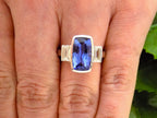 This is a 4.50ct total weight natural bezel set elongated cushion cut tanzanite ring with a single bezel set natural diamond on either side shown on a woman's finger.