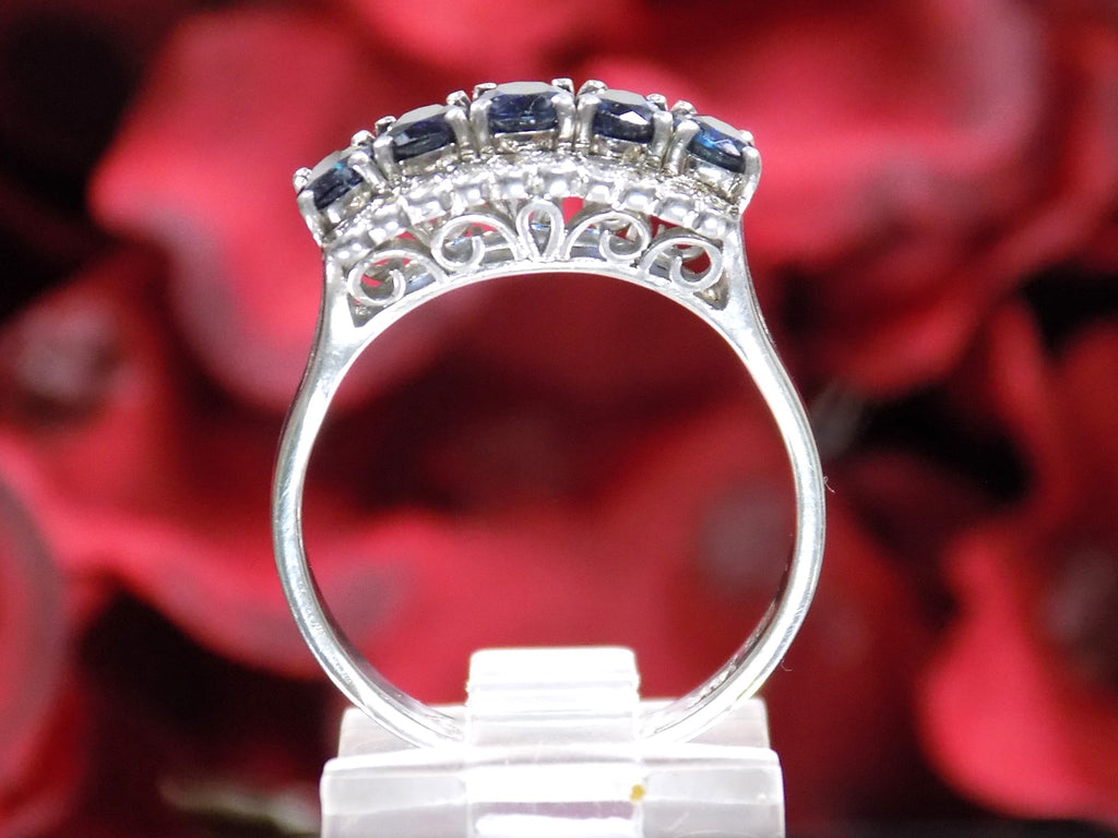 This is a women's vintage estate ring with a total of 2.00ct in natural oval sapphires and round diamonds in 14K white gold with a red floral background from a side view.