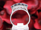 This is a women's vintage estate ring with a total of 2.00ct in natural oval sapphires and round diamonds in 14K white gold with a red floral background from a side view.
