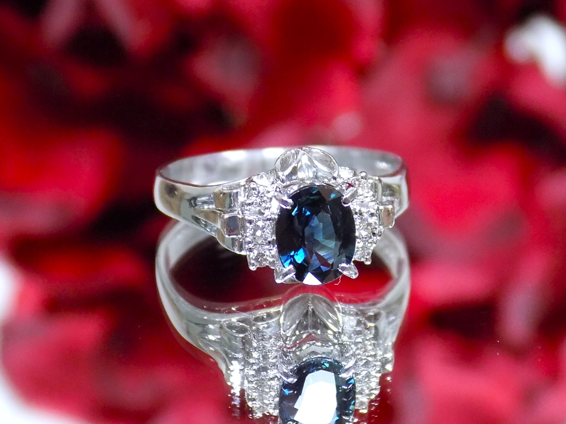 This is a women's vintage pre-world war II genuine oval shape sapphire and natural diamond ring 1.36ct total weight in platinum restored to showcase condition shown on a red flowered mirrored background.